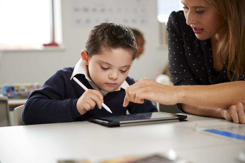 Young female teacher working with a Down syndrome schoolboy sitting at desk using a tablet computer in a primary school classroom, 前视图, 近距离