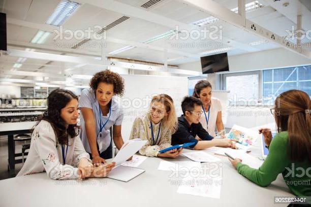 Teacher with a group of university students, in a laboratory classroom. The instructor is considering one of the students work, the mood is light hearted and positive. Other classmates are discussing things with each other. This is a realistic teaching scenario, with candid expressions. This is a multi-ethnic group of women. In the background there is a white board with mathematical formula written on it. All ladies are wearing id tags.