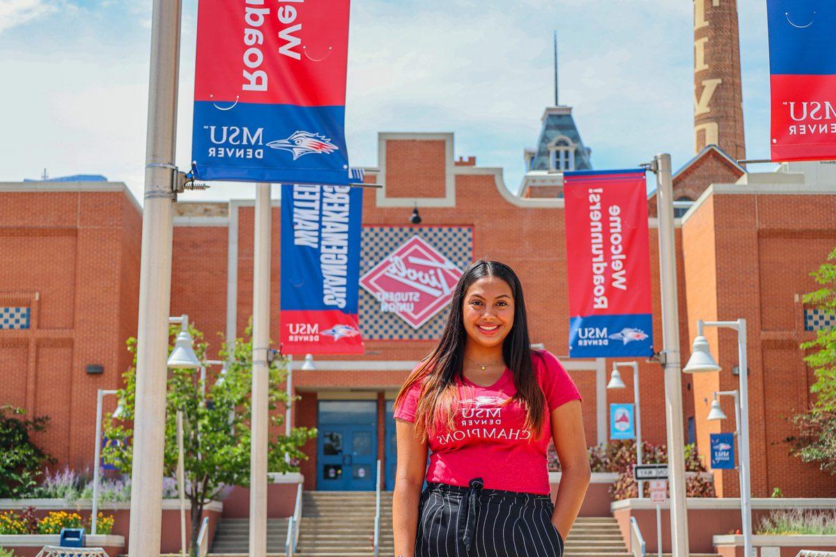 Student Ashley standing in front of the west side of the Tivoli with red and blue MSU Denver signs behind her