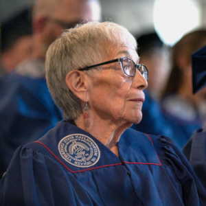 Photo of Anna Jo Garcia Haynes in MSU Denver regalia sitting watching the ceremony at the Spring 2023 Master of Arts in Teaching Hooding Ceremony.